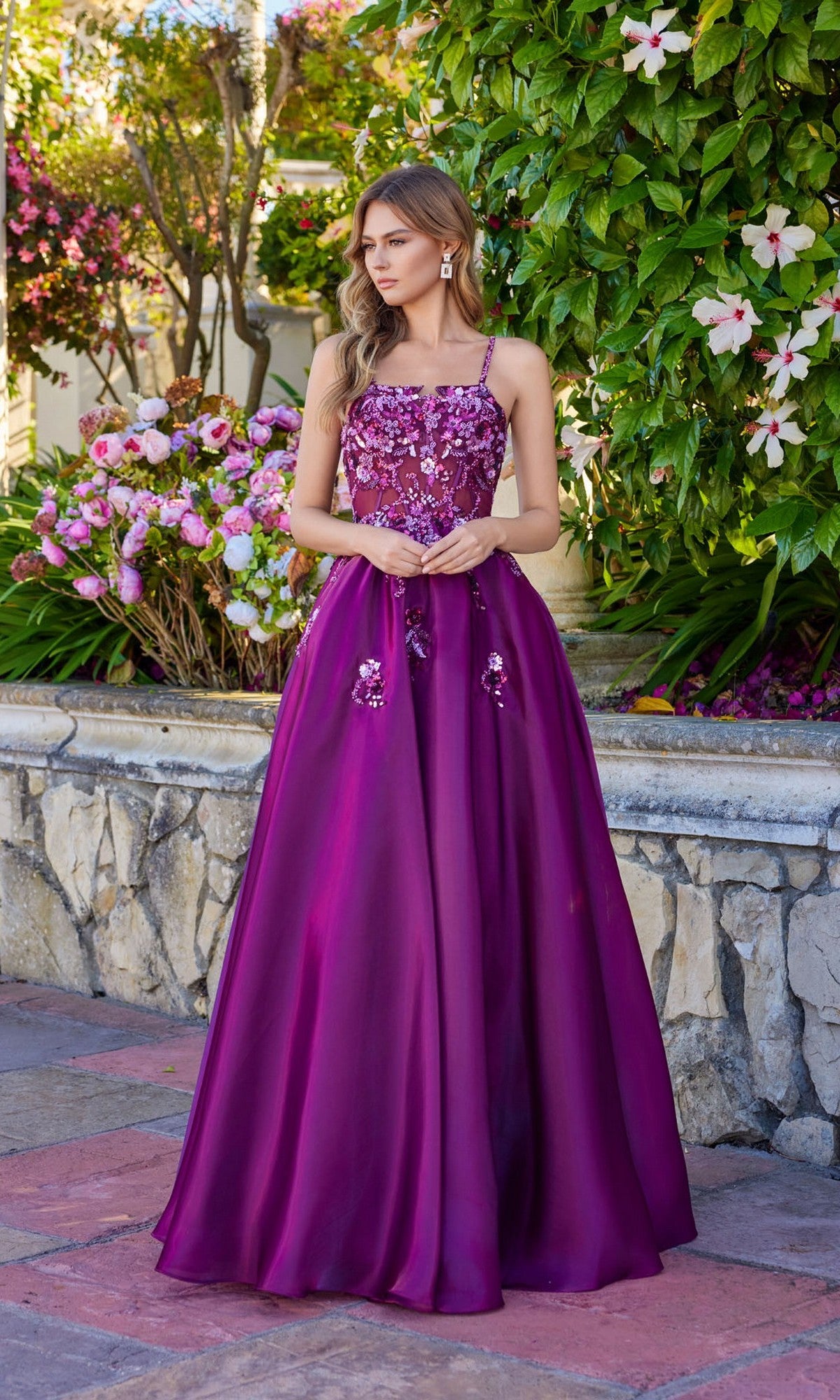 A woman stands outside by flowers and a stone wall, wearing the Juliet JT2617U long prom dress in wine, featuring a sleeveless A-line design with floral sequin embellishments on the bodice.